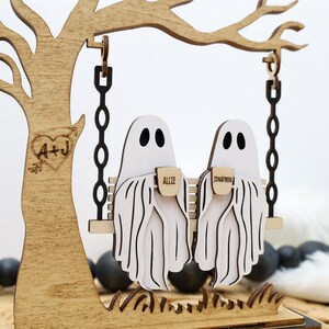 May include: Wooden Halloween decoration featuring two ghost figures on a swing. The ghosts are white with black eyes and are holding mugs with names on them. The swing hangs from a wooden tree with a heart and initials carved into it.