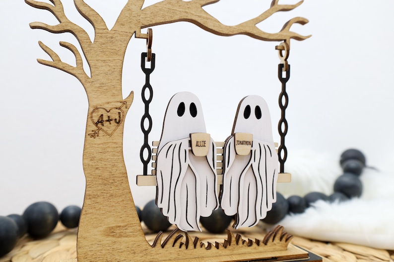 May include: Wooden Halloween decoration featuring two ghost figures on a swing. The ghosts are white with black eyes and are holding mugs with names on them. The swing hangs from a wooden tree with a heart and initials carved into it.