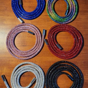 May include: Six coiled cords covered in small reflective crystals, each featuring metallic silver or black end caps. The cords are available in blue, a rainbow gradient, light pink, red, silver, and black, arranged in two columns of three.