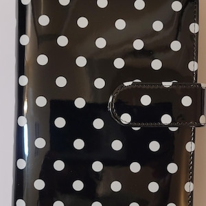 May include: A rectangular personal organizer with a glossy black finish and a repeating white polka dot pattern. The cover features a secure tab closure on the right side, maintaining a sleek, uniform appearance across the entire front surface.