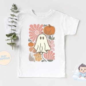 May include: White t-shirt with a retro design featuring a ghost, pumpkins, flowers, and spiderwebs. The design uses orange, peach, and cream colours, ideal for Halloween or autumn. The shirt is short-sleeved.