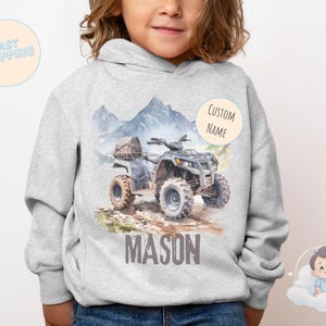 K&ouml;nnte beinhalten: Ein hellgrauer Kapuzenpullover mit einer Aquarellillustration eines blau-grauen ATVs auf einem Feldweg, mit Bergen im Hintergrund. Der Name "MASON" ist darunter aufgedruckt. Ein "CUSTOM NAME" Aufkleber befindet sich auf der rechten Seite.