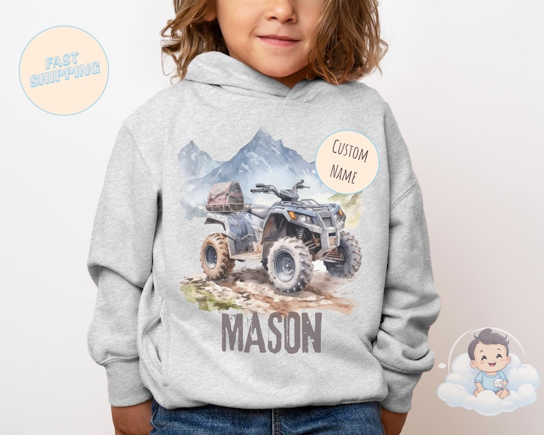K&ouml;nnte beinhalten: Ein hellgrauer Kapuzenpullover mit einer Aquarellillustration eines blau-grauen ATVs auf einem Feldweg, mit Bergen im Hintergrund. Der Name "MASON" ist darunter aufgedruckt. Ein "CUSTOM NAME" Aufkleber befindet sich auf der rechten Seite.