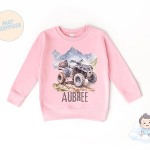 K&ouml;nnte beinhalten: Ein rosa Langarm-Sweatshirt mit einer Aquarellillustration eines ATVs auf einem Feldweg, mit Bergen im Hintergrund. Der Name "AUBREE" ist darunter aufgedruckt.