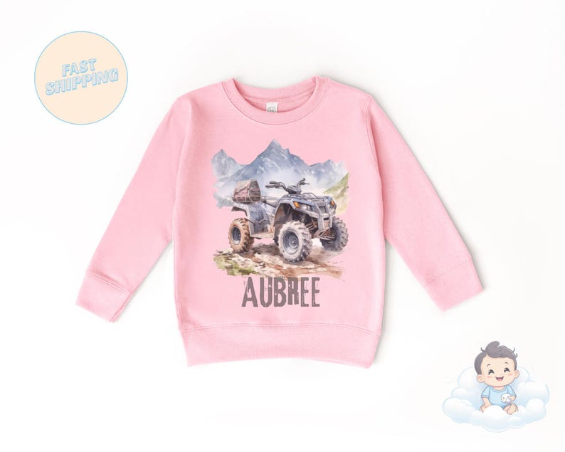 K&ouml;nnte beinhalten: Ein rosa Langarm-Sweatshirt mit einer Aquarellillustration eines ATVs auf einem Feldweg, mit Bergen im Hintergrund. Der Name "AUBREE" ist darunter aufgedruckt.