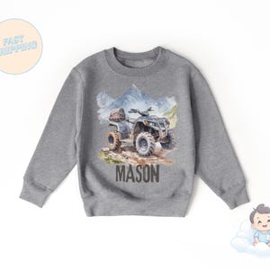 K&ouml;nnte beinhalten: Graues Sweatshirt mit Aquarell-Illustration eines ATVs und Berglandschaft. Der Name "MASON" ist in Braun unter dem ATV aufgedruckt. Das Sweatshirt hat einen Rundhalsausschnitt.