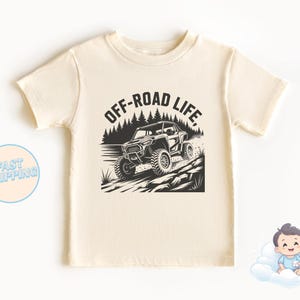 Könnte beinhalten: Cremefarbenes T-Shirt mit einer schwarz-weißen Grafik eines Geländewagens auf felsigem Gelände, mit dem Text "OFF-ROAD LIFE". Das Shirt hat kurze Ärmel und einen Rundhalsausschnitt.
