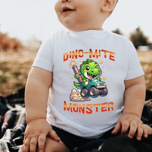 May include: A white t-shirt with a green dinosaur holding a firework and driving a monster truck. The text on the shirt reads "Dino-mite Monster".
