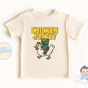 Puede incluir: Una camiseta beige con un personaje verde de Minecraft montando un pollo amarillo. El texto "Chicken Jockey" está impreso en amarillo en la camiseta.