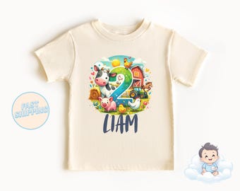 Puede incluir: Una camiseta beige claro con una escena de granja colorida que presenta una vaca, un cerdo, pollos y un tractor. El número 2 está en el centro del diseño. El texto "LIAM" está impreso debajo de la escena de la granja.
