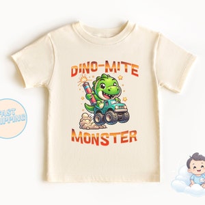 May include: A light beige t-shirt with a cartoon dinosaur driving a monster truck. The dinosaur is green and has a red firework in its mouth. The text "Dino-mite Monster" is printed in orange on the shirt.
