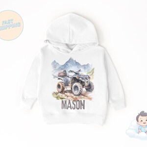 K&ouml;nnte beinhalten: Wei&szlig;er Kapuzenpullover mit einer Aquarellillustration eines Quads auf einem Feldweg, mit Bergen im Hintergrund. Der Name "MASON" ist in braunen Blockbuchstaben aufgedruckt. Der Pullover hat ein klassisches Design.