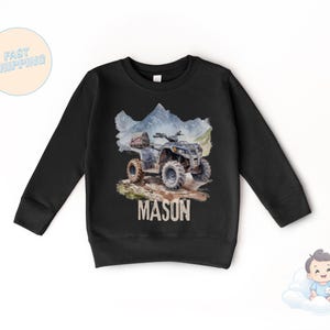 K&ouml;nnte beinhalten: Schwarzes Langarm-Sweatshirt mit einer Aquarell-Illustration eines ATVs auf einem schlammigen Pfad und Bergen. Der Name "MASON" ist darunter gedruckt. Das Sweatshirt ist f&uuml;r Kinder.
