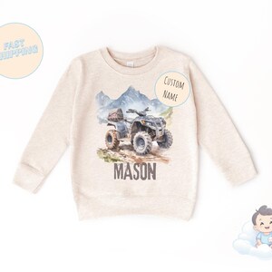 K&ouml;nnte beinhalten: Ein beiges Sweatshirt mit einer Aquarellillustration eines ATVs auf einem Bergpfad. Der Name "MASON" ist unter dem ATV aufgedruckt. Die Worte "CUSTOM NAME" stehen auf einem Aufkleber. Die Worte "FAST SHIPPING" stehen auf einem Aufkleber.