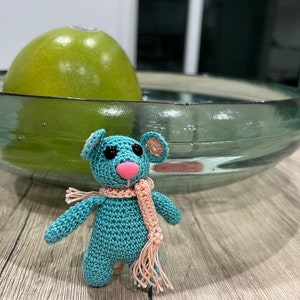 Puede incluir: Un pequeño ratón de crochet de color turquesa con una bufanda rosa. El ratón tiene ojos negros y una nariz rosa.