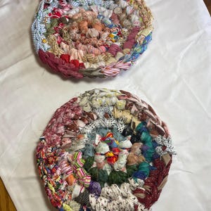 May include: Two handmade, circular trivets crafted from colorful fabric scraps. The trivets feature a spiral design with a variety of patterns and hues, including red, blue, pink, and brown. These trivets are a charming addition to any kitchen.