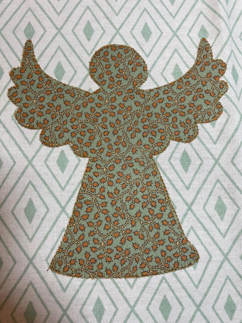 May include: A fabric angel applique with outstretched wings, featuring a green and brown floral pattern. The angel is set against a white background with a diamond pattern in light green. This decorative piece is suitable for crafts and home decor.
