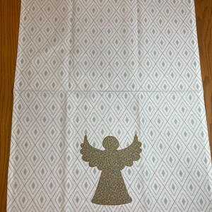 May include: White and gray patterned decorative towel featuring a diamond design. The towel has a brown angel applique with a speckled pattern. The towel is made of a soft material and is suitable for kitchen or bathroom use.