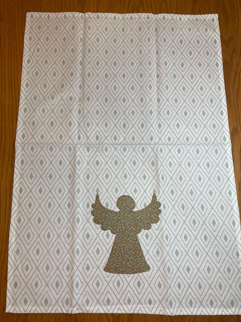 May include: White and gray patterned decorative towel featuring a diamond design. The towel has a brown angel applique with a speckled pattern. The towel is made of a soft material and is suitable for kitchen or bathroom use.