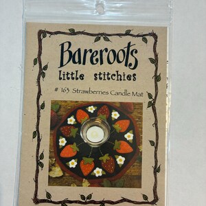Puede incluir: Un paquete para un tapete de velas con diseño de fresas. El paquete está etiquetado como "Bareroots little stitches" y presenta un tapete circular con fresas, flores blancas y un portavelas en el centro.