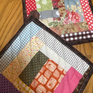 Puede incluir: Dos salvamanteles acolchados hechos a mano con un diseño de patchwork. Uno presenta estampados florales, de lunares y cuadros vichy en rojo, azul y marrón. El otro tiene una variedad de patrones geométricos coloridos, incluyendo lunares y cuadros.