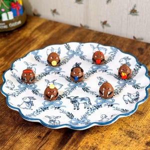 May include: A decorative white plate with a blue floral pattern and scalloped edges holds eight chocolate egg-shaped candies, each with a unique cartoon face. The plate sits on a wooden surface.