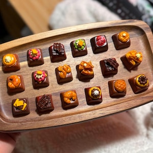 May include: A wooden tray with 18 small square keycaps featuring various food designs, including pancakes, waffles, croissants, and donuts. The keycaps are arranged in a grid pattern on the tray.