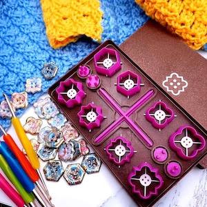 Puede incluir: Una herramienta de plástico marrón con moldes rosas para hacer diferentes formas de botones. La herramienta tiene un diseño en forma de cruz con dos brazos largos y dos brazos cortos. Los moldes están dispuestos en una cuadrícula. También hay dos moldes redondos pequeños con clavijas en el centro. La herramienta está rodeada de una variedad de botones coloridos.