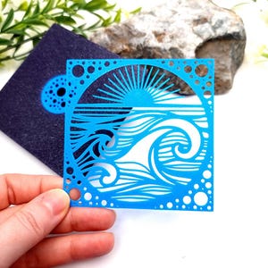 May include: A vibrant blue paper cut-out depicting a sun setting over ocean waves. The design includes a square border with circular cut-outs. A dark blue square with a circular design is in the background.