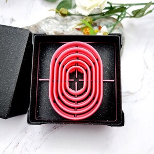 May include: A pink, oval-shaped cookie cutter set nested in a black box. The set includes multiple sizes of the same shape, perfect for baking. The cutter is made of plastic and sits on a black surface.