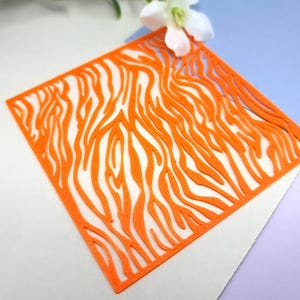 May include: Orange paper stencil with a zebra stripe pattern. The stencil is cut out in the shape of a square.