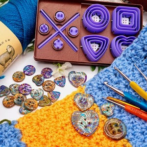 May include: A brown plastic organizer tray containing purple circular, square, triangular, and heart-shaped loom tools, alongside a collection of multicolored patterned buttons, three metal crochet hooks, and a blue and yellow crocheted fabric.