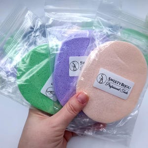 May include: Three makeup sponges in individual plastic bags. The sponges are in the colors green, purple, and peach. The bags have the text "Sweety Bijou Professional Tools" printed on them.