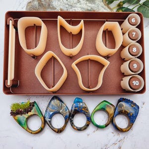 May include: A collection of handmade rings and jewelry-making tools. The rings feature unique designs with blue, green, and gold resin. The tools include various shaped cutters and cylindrical molds with numbers 6-10, all in a brown tray.