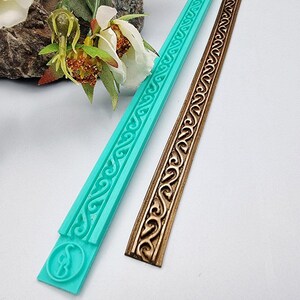 May include: A teal silicone mould and a brown decorative trim featuring an ornate, repeating scroll design. The mould has a circular emblem with a stylized letter "B". The trim is a finished product, while the teal item is the mould used to create it.