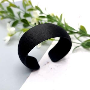 May include: A black, wide, open-ended bangle bracelet made of metal. The bracelet is smooth and has a matte finish.