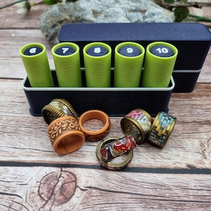May include: A dark blue storage box containing five lime green cylinders labelled with the numbers 6 through 10. In front of the box are several rings, including one with a floral pattern, one with a mosaic tile design, and one with a red swirl.