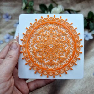 May include: A square white tile featuring a raised, bright orange mandala design. The intricate pattern consists of concentric circles, petal-like shapes, and small teardrop accents radiating from the center to the outer scalloped edge.