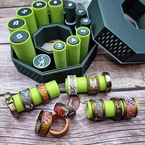May include: A dark green octagonal organizer containing multiple light green cylindrical ring holders, each marked with a number. Several decorative rings with varied textures, metallic finishes, and colorful mosaic patterns are displayed.