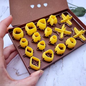 May include: A collection of yellow polymer clay cutters in various shapes, including circles, squares, and flower-like designs, displayed in a brown box. The cutters are arranged on a brown tray, showcasing their different forms. The box is open, revealing the contents.