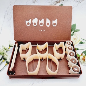 Puede incluir: Una caja metálica marrón con un juego de cortadores de galletas beige e insertos marrones numerados. La tapa de la caja tiene un diseño blanco. Los cortadores de galletas tienen varias formas. Los insertos están numerados del 6 al 10.