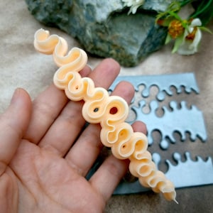 May include: A pale peach-colored, wavy, serpentine-shaped plastic tool with a continuous, undulating edge. The tool features a series of interconnected, rounded loops and curves, designed for creating decorative icing patterns or textured clay designs.