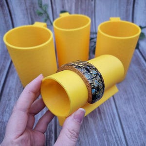 May include: A yellow cylindrical object with a handle, holding a decorative bracelet. The bracelet has a brown leather band and a mosaic design with black, gold, and turquoise elements. Three other yellow cylinders are in the background.