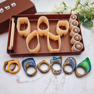 May include: A collection of handmade rings and ring sizers. The rings feature unique designs with blue, green, and gold resin, and wood. The ring sizers are beige plastic and numbered 6-10. A brown tray holds the ring sizers and ring molds.