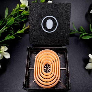 May include: A black square box with a white oval design on top, and a set of orange oval-shaped molds inside a black container. The molds are nested within each other, with a star-like design in the center. The background is black with white floral accents.