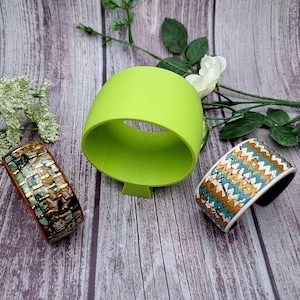 May include: Three bracelets and a lime green cylindrical object are displayed on a wooden surface. The bracelets feature intricate patterns in gold, teal, and white. The green object has a circular opening.