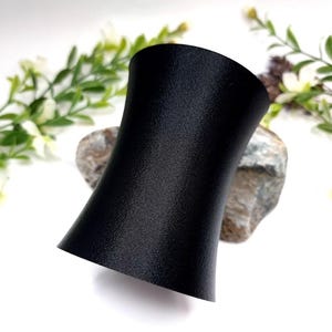 May include: A black, wide, adjustable bracelet with a smooth, matte finish. The bracelet is made of a durable material and has a slightly curved shape.