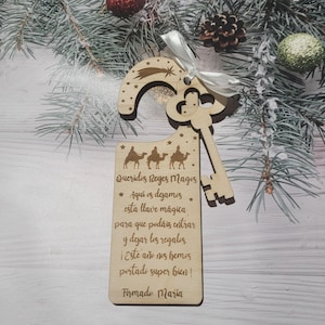 Könnte beinhalten: Ein hölzerner Weihnachts-Türhänger in Form eines Schlüssels mit einem Kometen- und Sterndesign. Der Anhänger hat Text auf Spanisch und ist mit einem weißen Band gebunden. Das Design zeigt drei Kamele und Reiter.