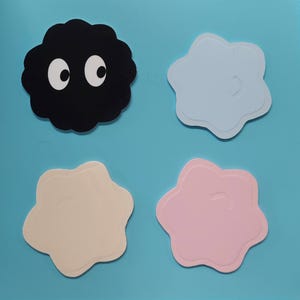 May include: Four coasters on a blue surface. One black coaster with white eyes, and three flower-shaped coasters in light blue, beige, and pink. The coasters have a layered design.