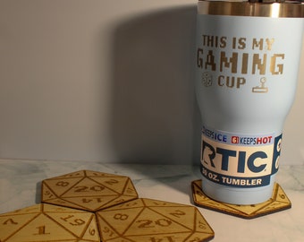 May include: A light blue 30 oz. RTIC tumbler with the text THIS IS MY GAMING CUP and icons of a die and joystick. Three wooden coasters shaped like 20-sided dice with engraved numbers are arranged around the base of the tumbler on a surface.